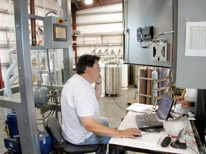 man on the phone at a desk with an open laptop, with various gas cylinders in the back ground